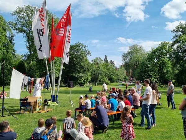 Auf diesem Bild sehen Sie Messebesucher/innen, die bei strahlendem Sonnenschein einem Kinderprogramm zuhören.